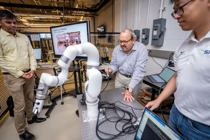 Three individuals are observing and discussing a robotic arm in a laboratory setting.