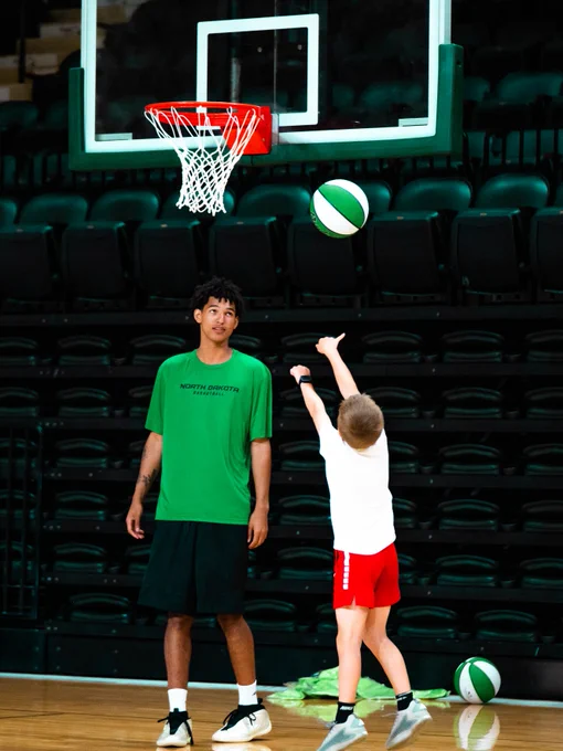 A student-athlete in a green shirt watches another in red shorts taking a basketball shot in a gym. A basketball hoop and two green and white basketballs are visible.