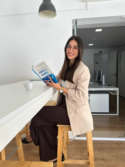A woman with long dark hair and glasses sits on a wooden stool at a high white table in a bright office space. She is smiling while holding a book in a beige blazer, white blouse, and dark trousers, she sits beside a white coffee mug. The modern office features white walls, wooden flooring, and workstations in the background.