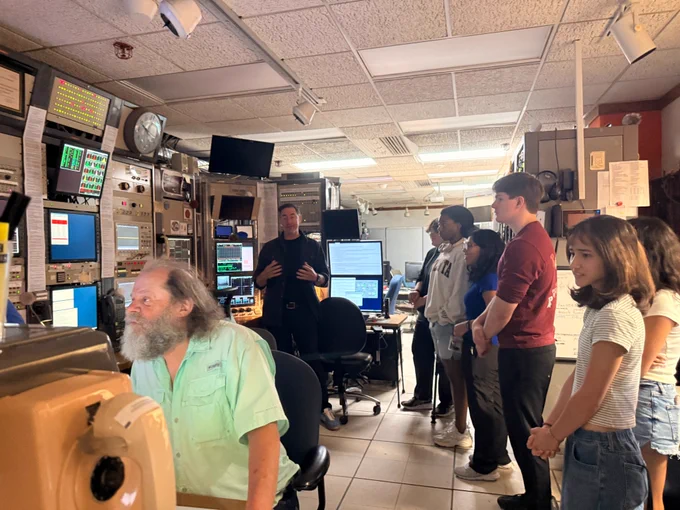 A tour guide speaks to a group of students gathered inside a computer-filled control room, with an operator seated at a control panel monitoring multiple screens.
