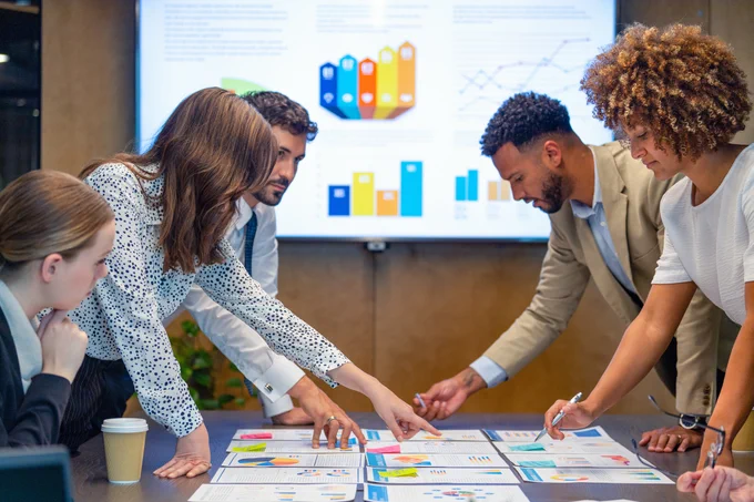 A group of people in professional attire are gathered around a conference table reviewing charts and documents. A large digital screen in the background displays colorful graphs and charts.