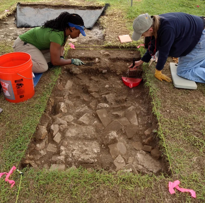 Two people kneeling by a rectangular archaeological excavation site, using brushes to clear dirt from stones.