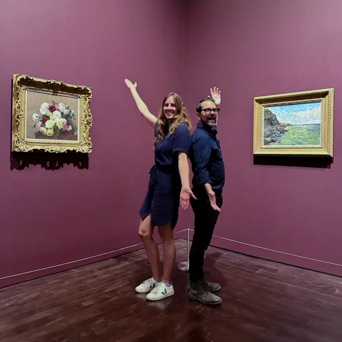 Two people stand back-to-back in a gallery with purple walls, smiling and gesturing towards two paintings. One painting features a bouquet of flowers, and the other depicts a coastal landscape.