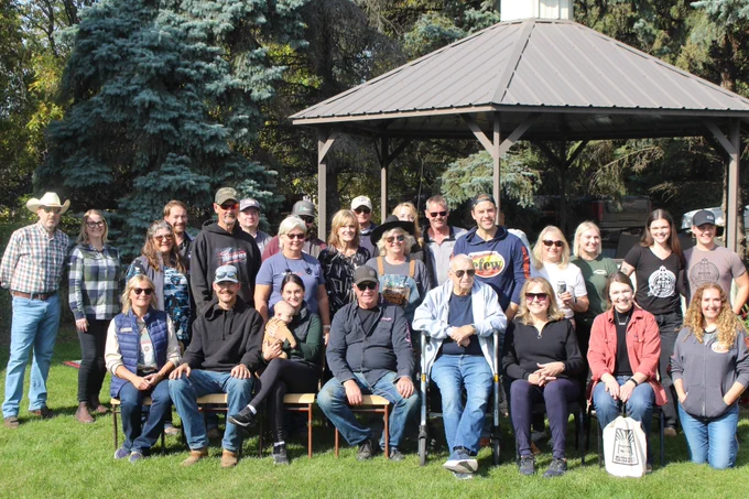 Jordyn Prior, family and friends, pose for a photo in an outdoor setting with trees and a gazebo. Some are seated while others stand behind them. They are wearing casual clothes, and the sun is shining brightly.
